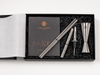 A silver metal jigger and strainer set with a black presentation box, shown on a white background. The set includes a double jigger, a pourer, and two strainers, arranged in black shredded paper next to a black card with gold logo and text that reads 'RUSTY BARREL'.