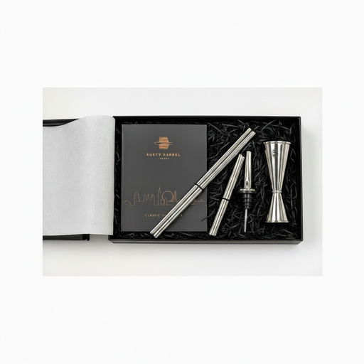 A silver metal jigger and strainer set with a black presentation box, shown on a white background. The set includes a double jigger, a pourer, and two strainers, arranged in black shredded paper next to a black card with gold logo and text that reads 'RUSTY BARREL'.