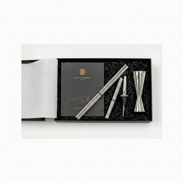 A silver metal jigger and strainer set with a black presentation box, shown on a white background. The set includes a double jigger, a pourer, and two strainers, arranged in black shredded paper next to a black card with gold logo and text that reads 'RUSTY BARREL'.