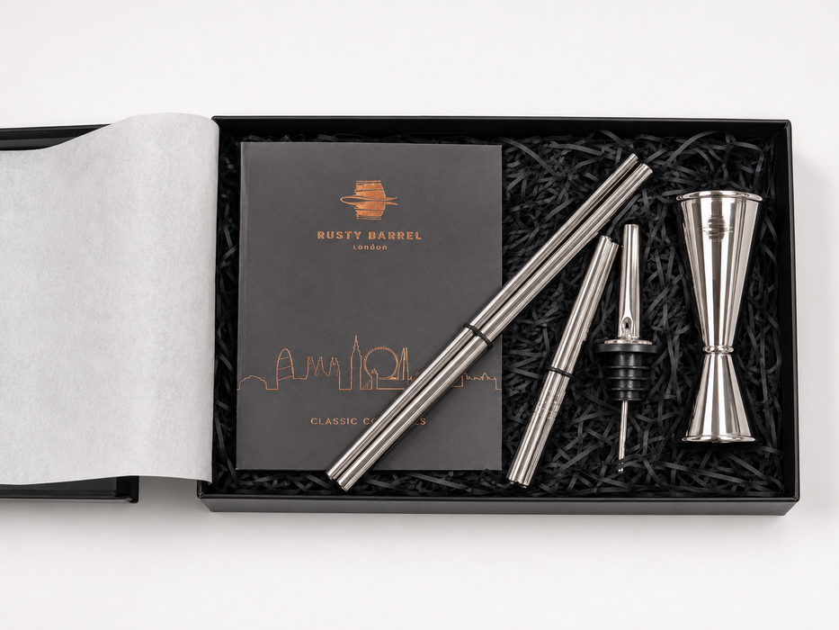 A silver metal jigger and strainer set with a black presentation box, shown on a white background. The set includes a double jigger, a pourer, and two strainers, arranged in black shredded paper next to a black card with gold logo and text that reads 'RUSTY BARREL'.